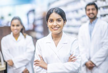 A diverse multi ethnic pharmacy team pose for a group portrait, and are lead by a female pharmacist.  A male pharmacists is out of focus behind her to the right and another female pharmacists is standing behind her out of focus to the left.  They are all smiling as they take great pride in their job as health care providers.